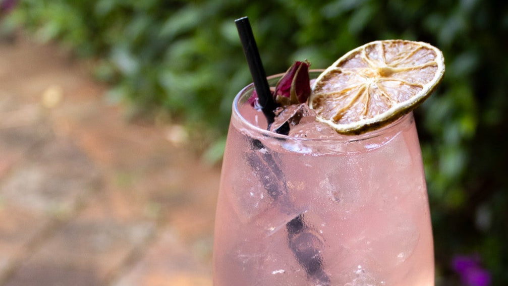 Pink cocktail with a lime wedge and straw on a wooden surface with greenery in the background