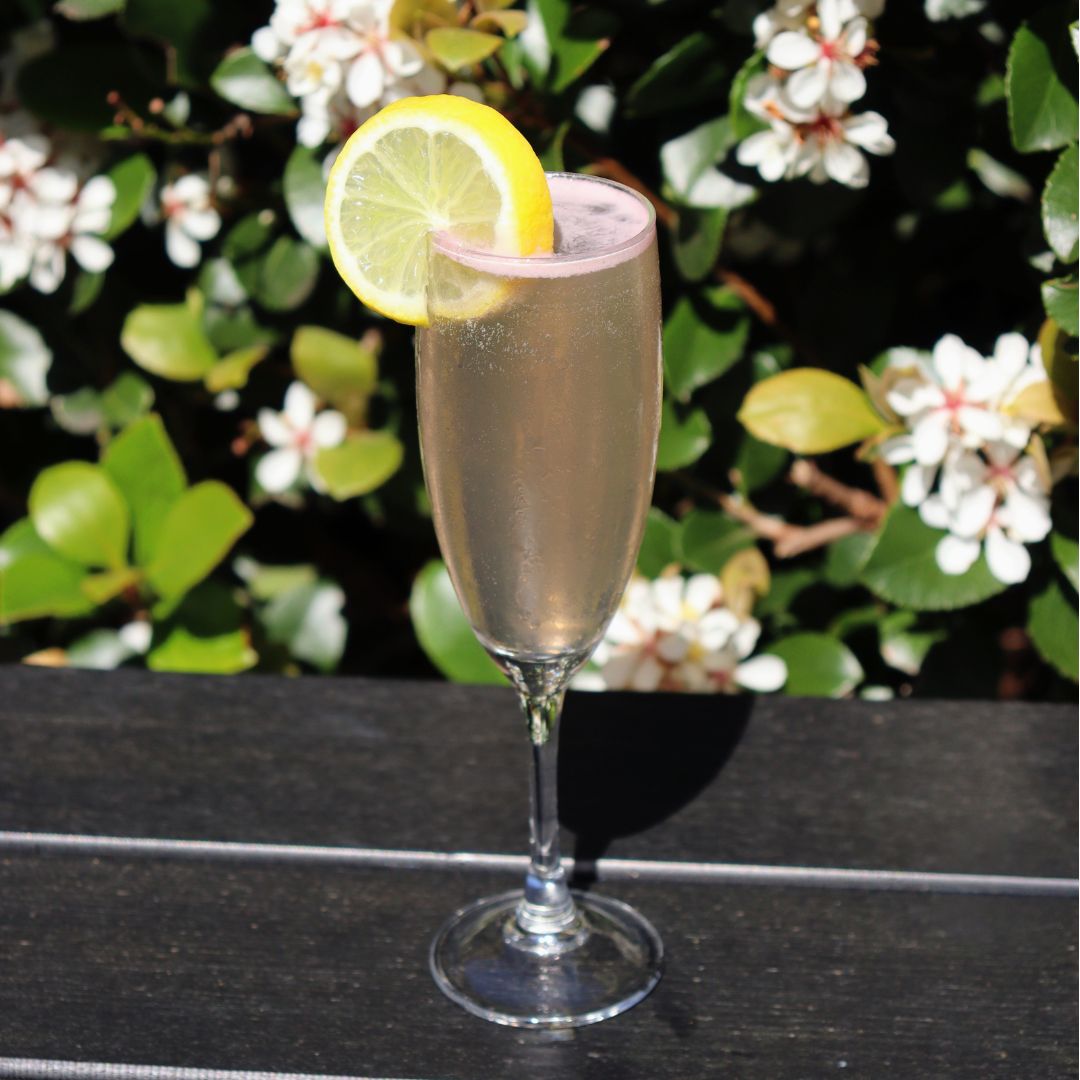 Flute glass with Violet 75 cocktail made with gin, violet liqueur, lemon juice and sparkling wine, garnished with lemon twist.